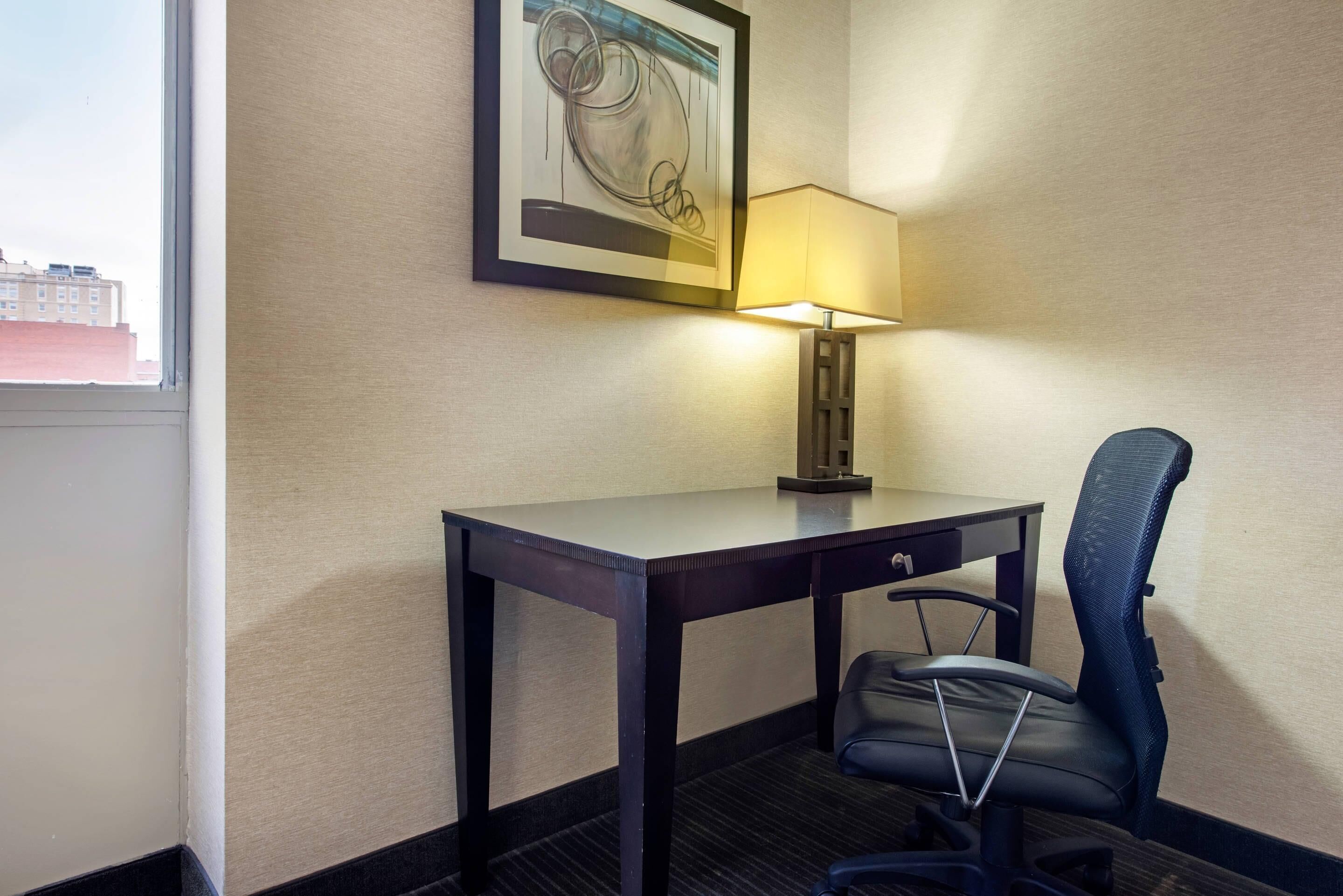 Habitación de hotel con escritorio, silla, lámpara y una obra de arte enmarcada en la pared.