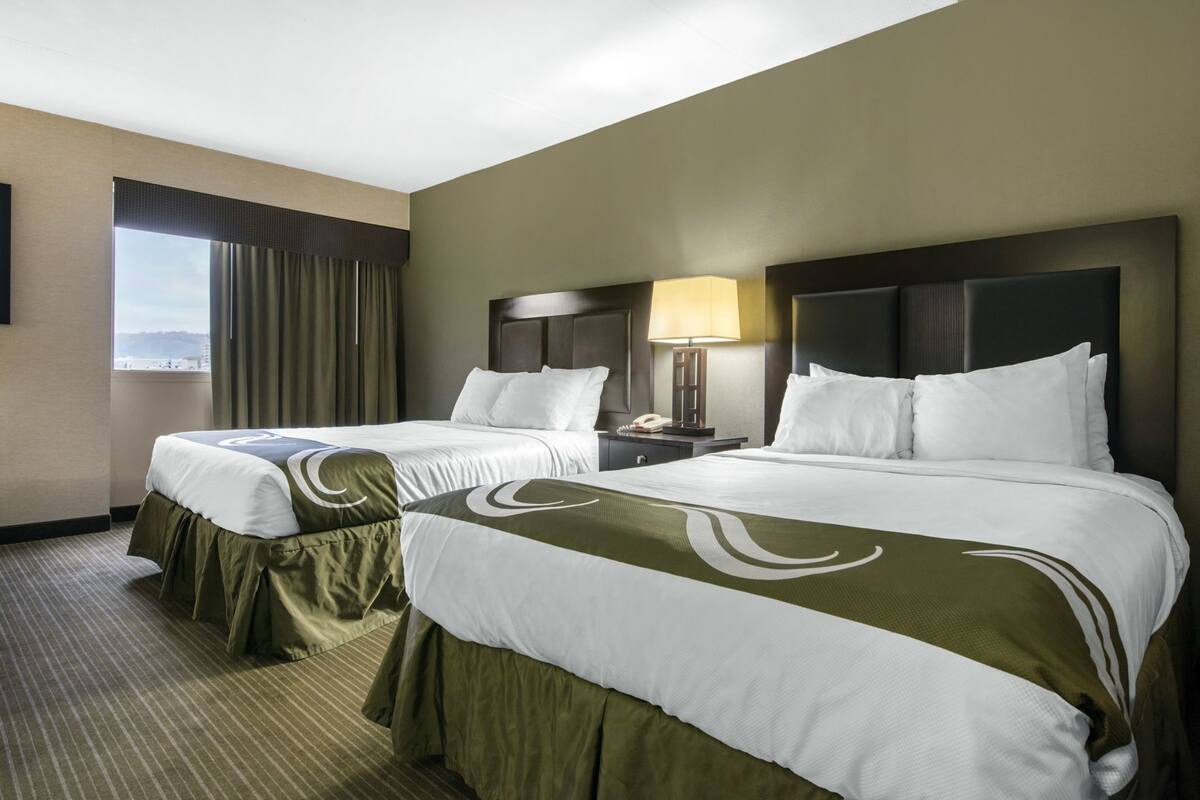 A hotel room with two beds, a window with curtains, a bedside table with a lamp, and a view of the outside.