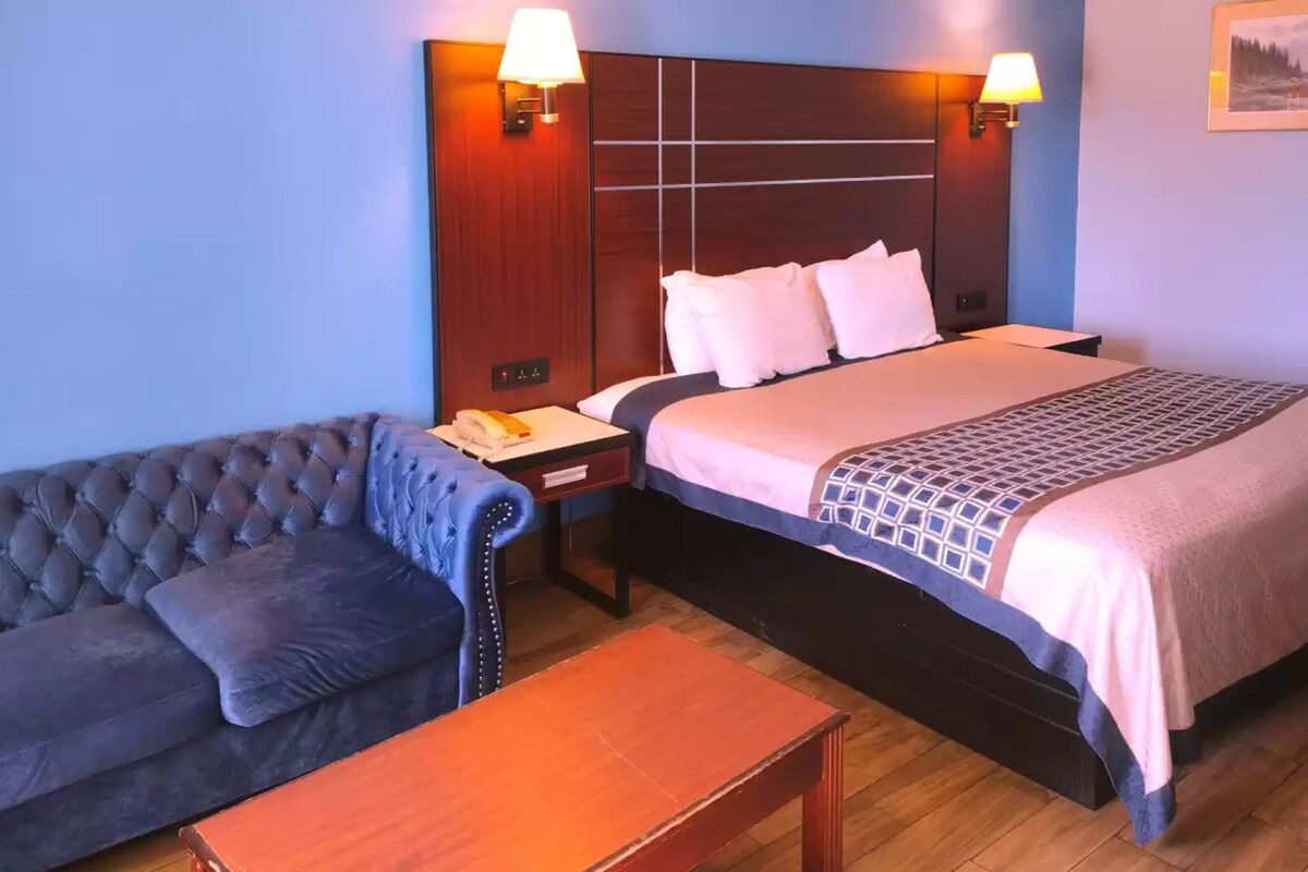 Una habitación de hotel con una cama grande, un sofá capitonado, una mesita de noche de madera y dos lámparas fijadas a la pared.