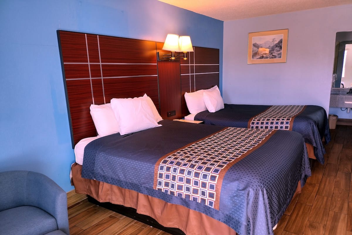 Habitación de hotel con dos camas, cabecera de madera, mesita de noche, lámpara, un cuadro en la pared y una silla.