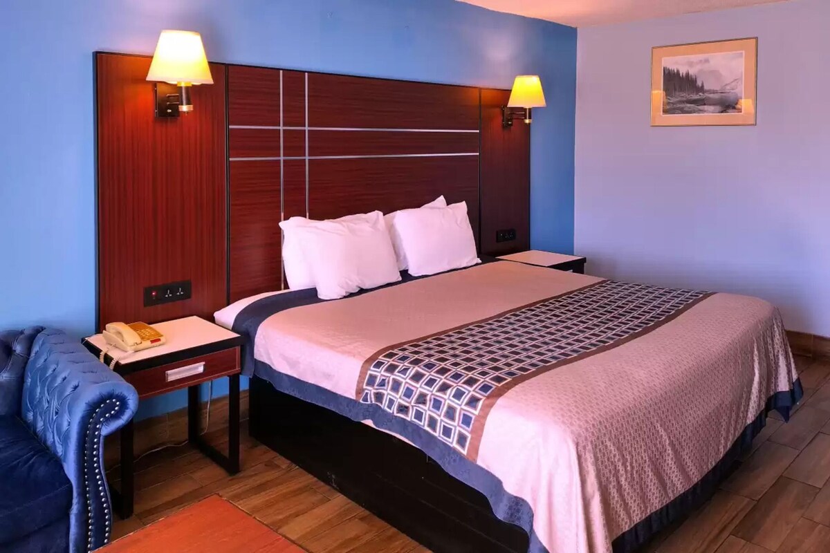 Una cama bien hecha con una colcha estampada, una mesita de noche con una lámpara y un sillón tapizado en azul.