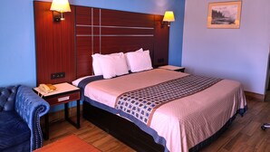 A hotel room with a large bed, two bedside lamps, a wooden headboard, a small table with a telephone, a blue armchair, and a framed picture on the wall. - Rodeway Inn Richland - Jackson South (Richland)