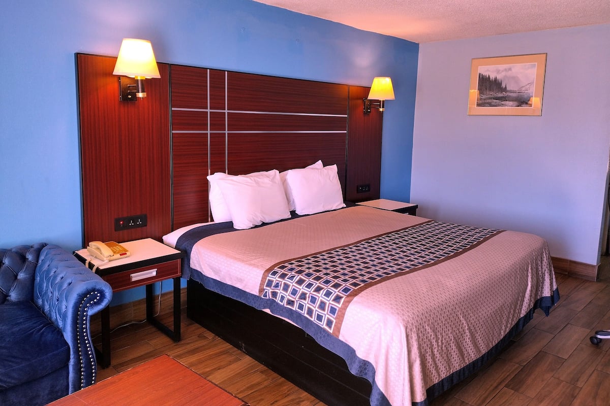 Habitación de hotel con una cama grande, dos lámparas de noche, un cabecero de madera, una mesita con un teléfono, un sillón azul y un cuadro en la pared.