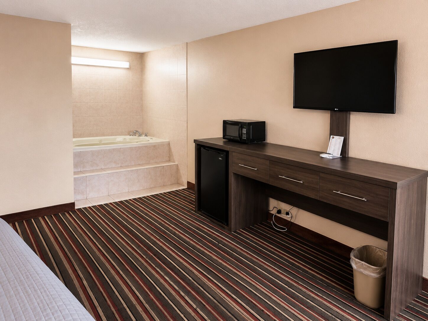 A hotel room with a bed, a small kitchenette, a flat-screen TV, a microwave, a trash bin, and a bathroom with a bathtub and stairs.