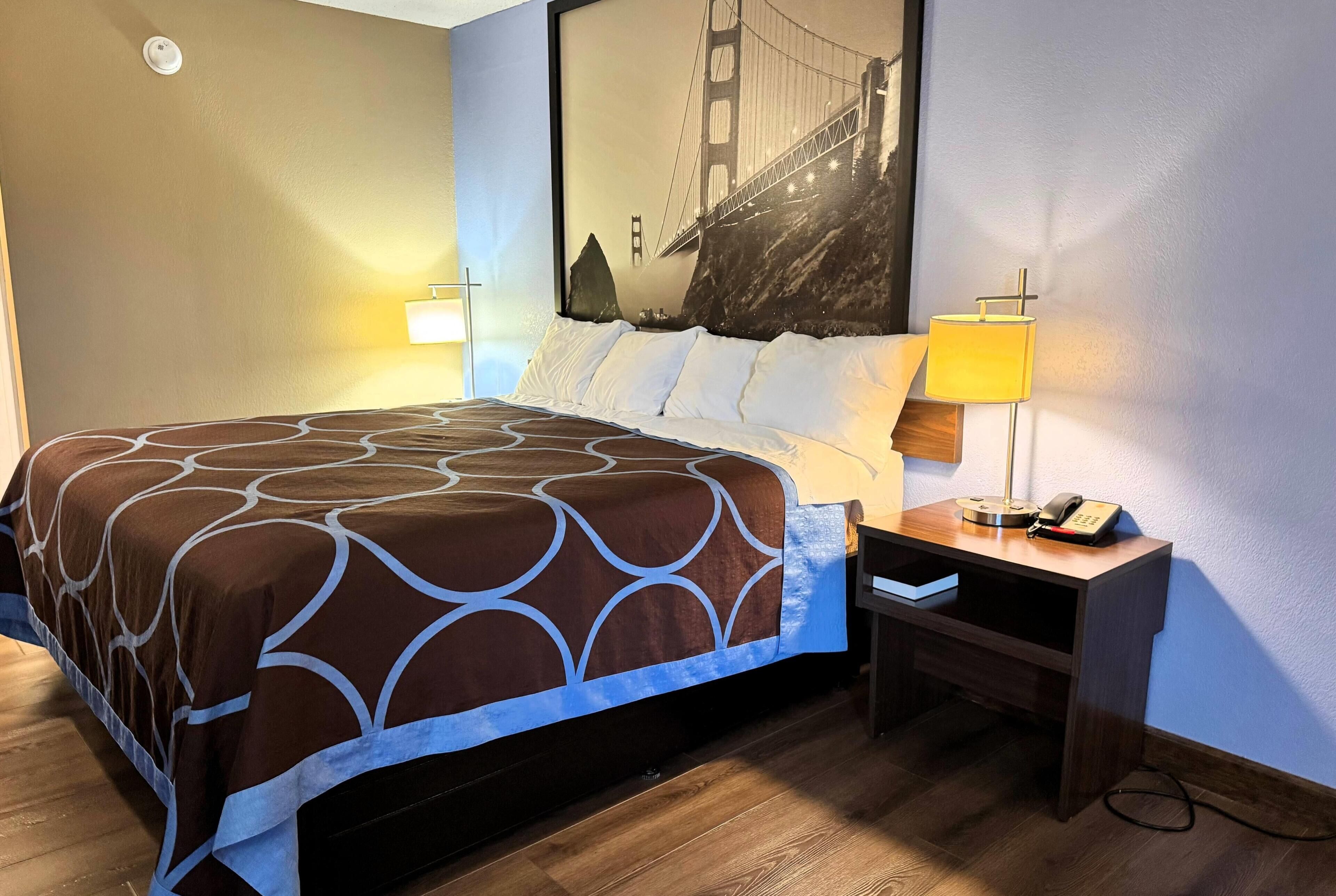A hotel room with a bed, two bedside lamps, a nightstand with a phone, and a large wall painting of the Golden Gate Bridge.