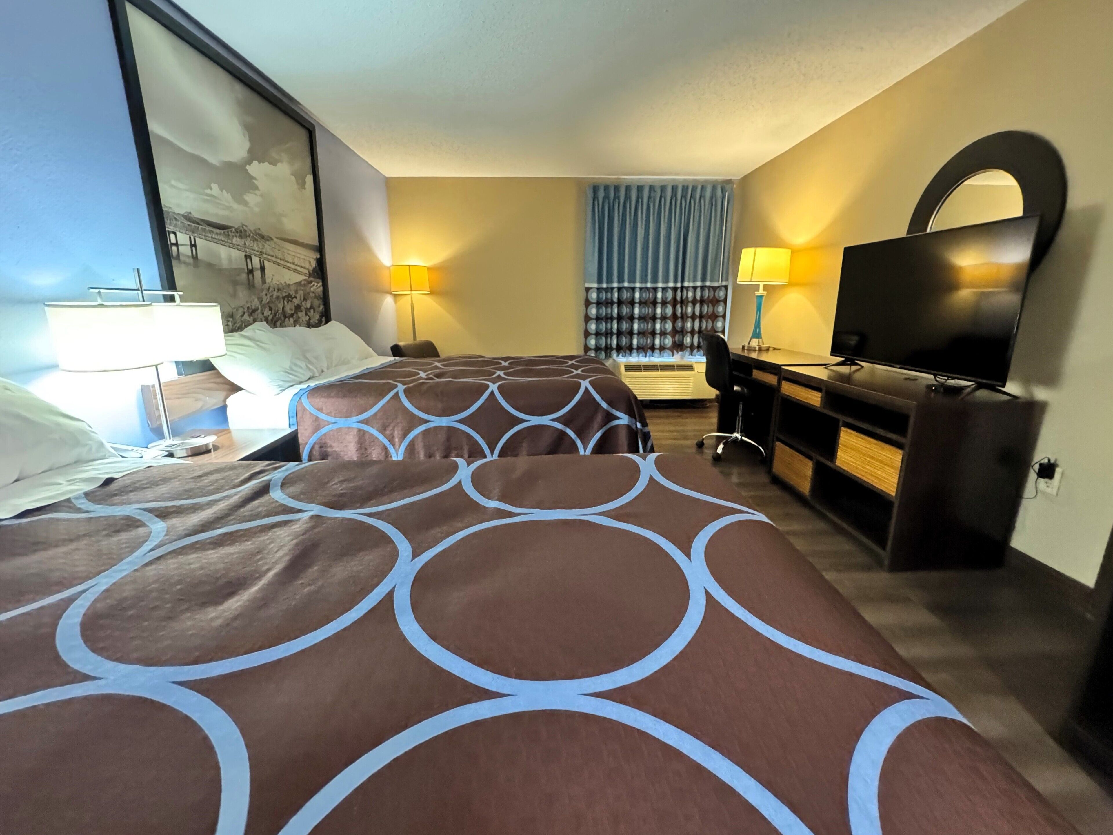 A hotel room with two beds, a desk, a TV, and a large window with curtains.