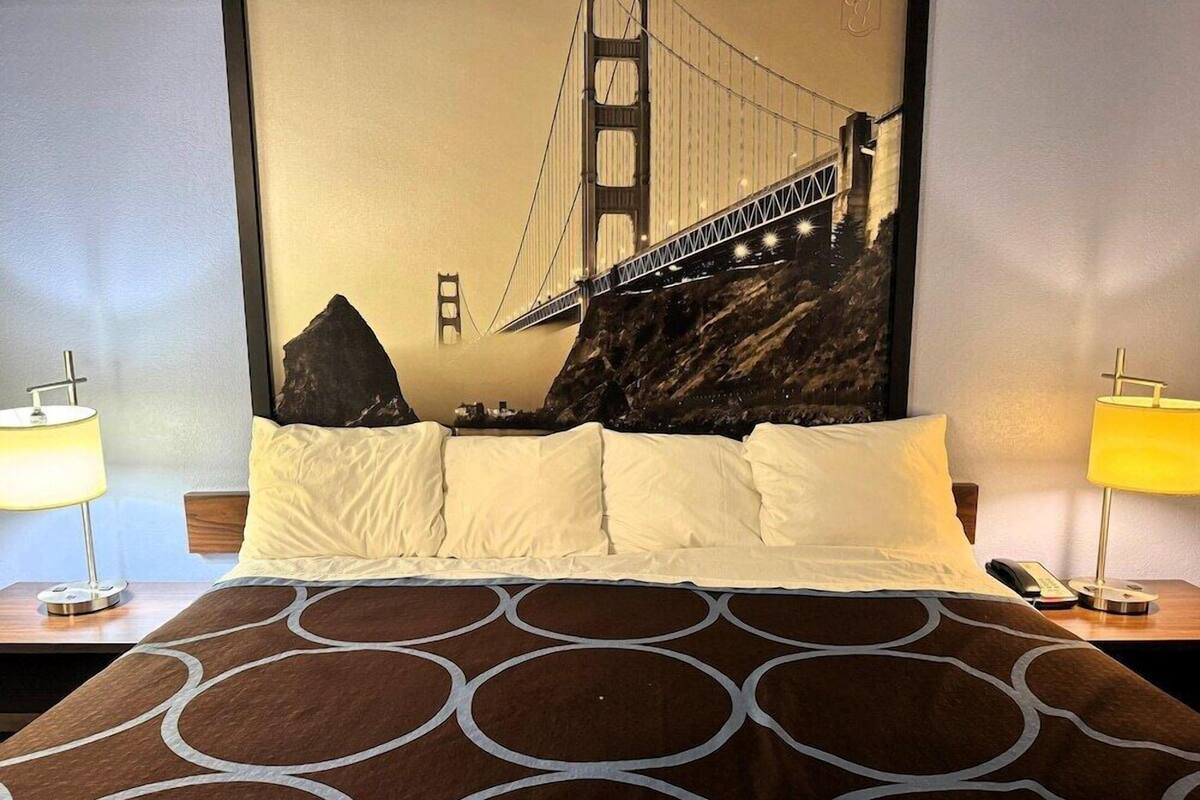 A hotel room with a bed, two bedside lamps, a wall-mounted telephone, and a large mural of the Golden Gate Bridge.