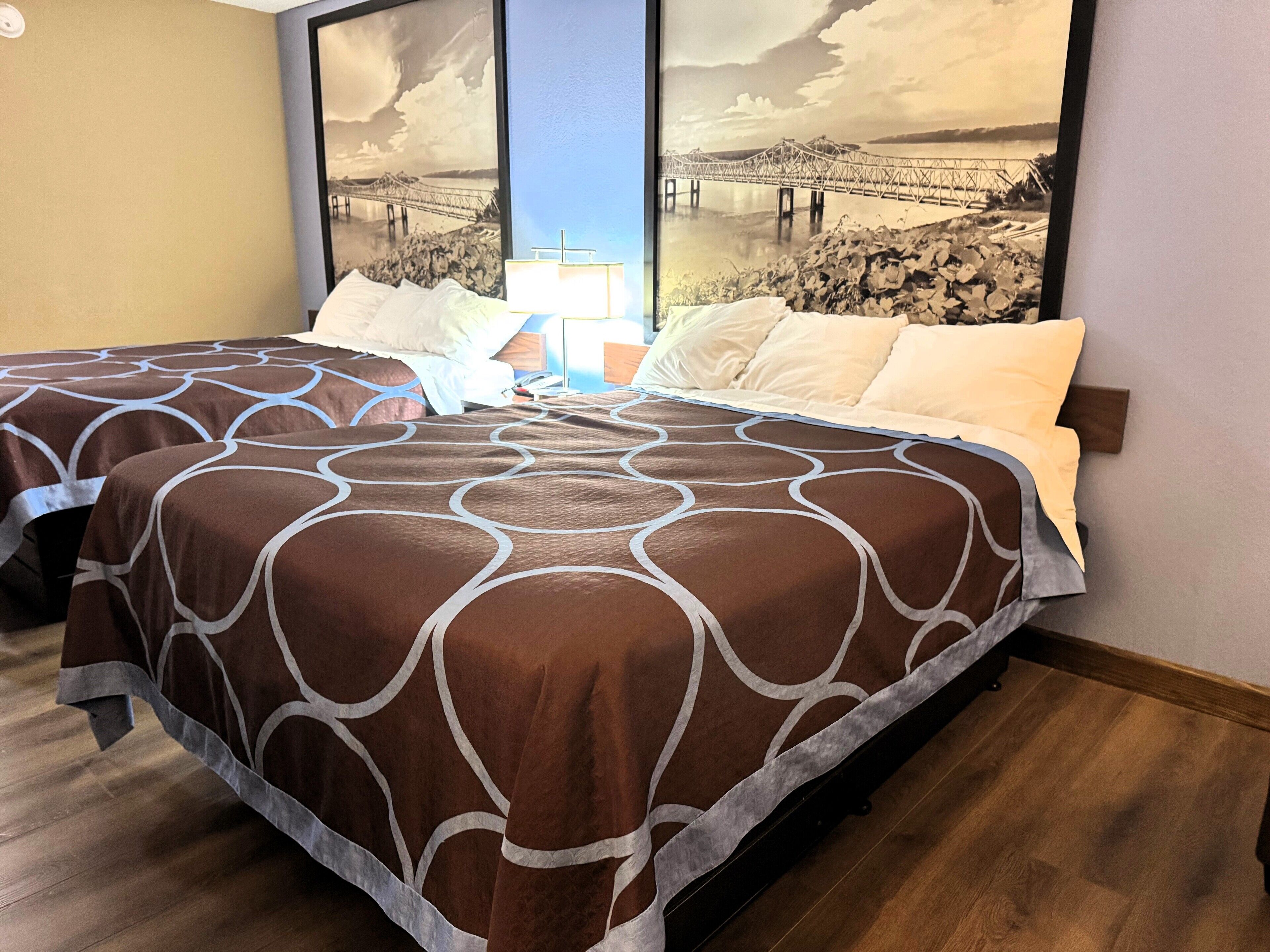 A hotel room with two beds, each with a brown and white patterned bedspread, a large window with a view of a bridge, and a wooden floor.