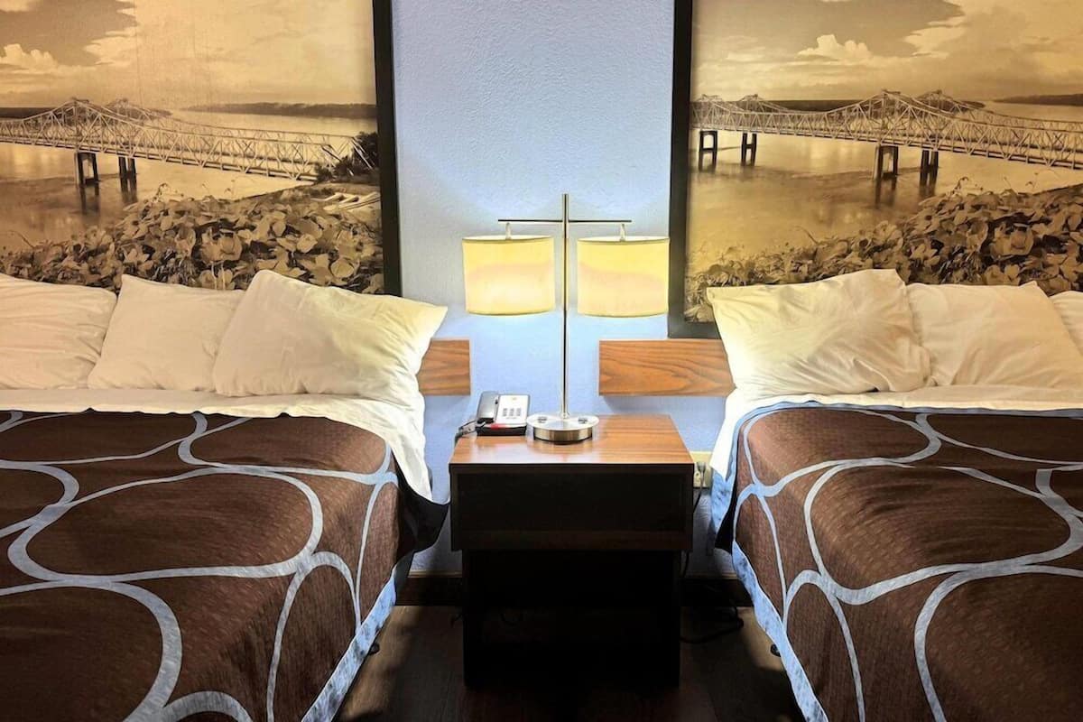 A hotel room with two beds, a nightstand with a lamp, and a large wall mural of a bridge over water.