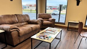 Una habitación con un sofá de cuero marrón, un sillón a juego, una mesa de centro de madera con revistas, y vistas a una gasolinera a través de amplios ventanales.