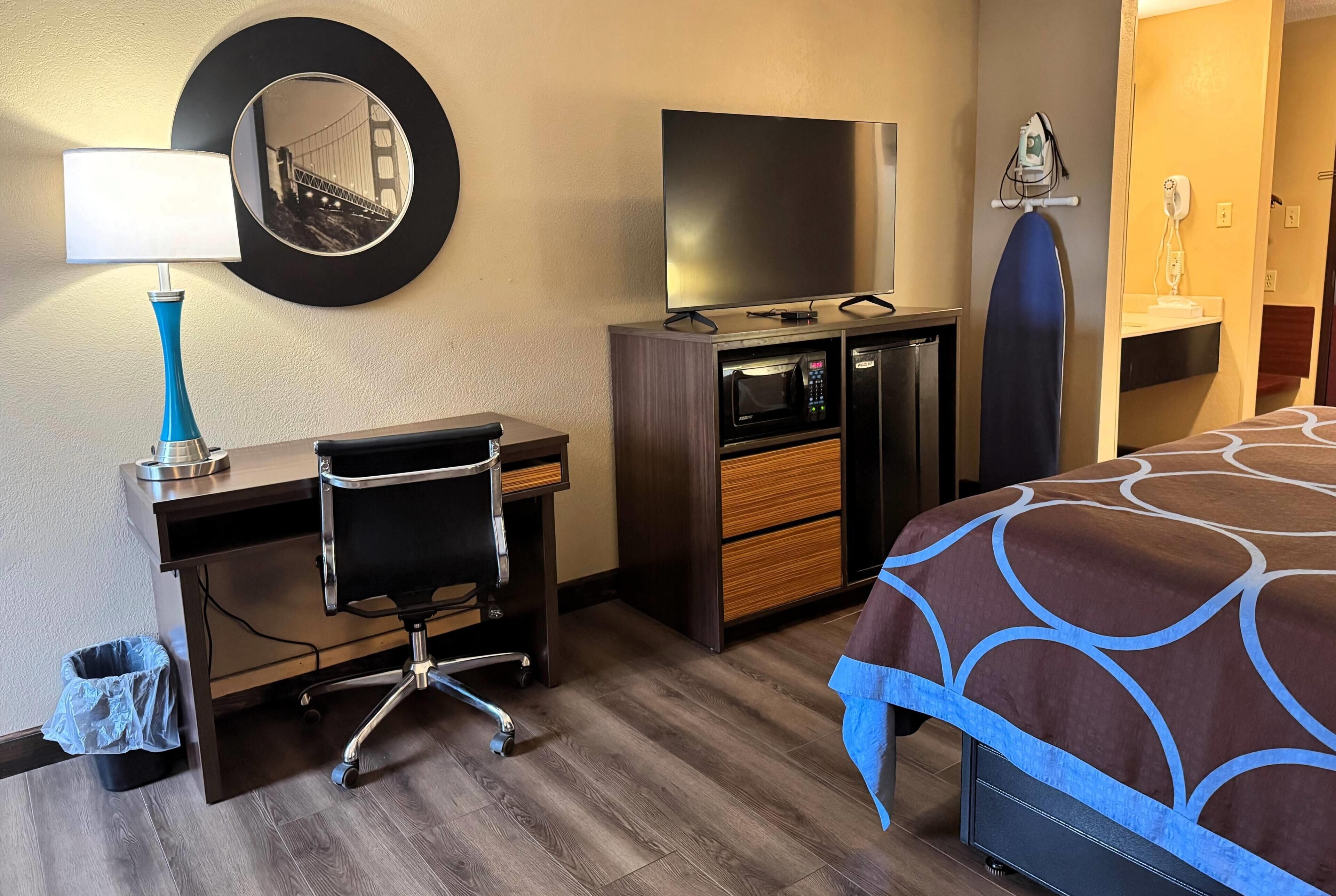 A hotel room with a desk, chair, TV, microwave, ironing board, and a bed with a patterned bedspread.