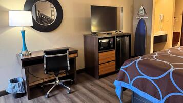 A hotel room with a desk, chair, TV, microwave, ironing board, and a bed with a patterned bedspread.