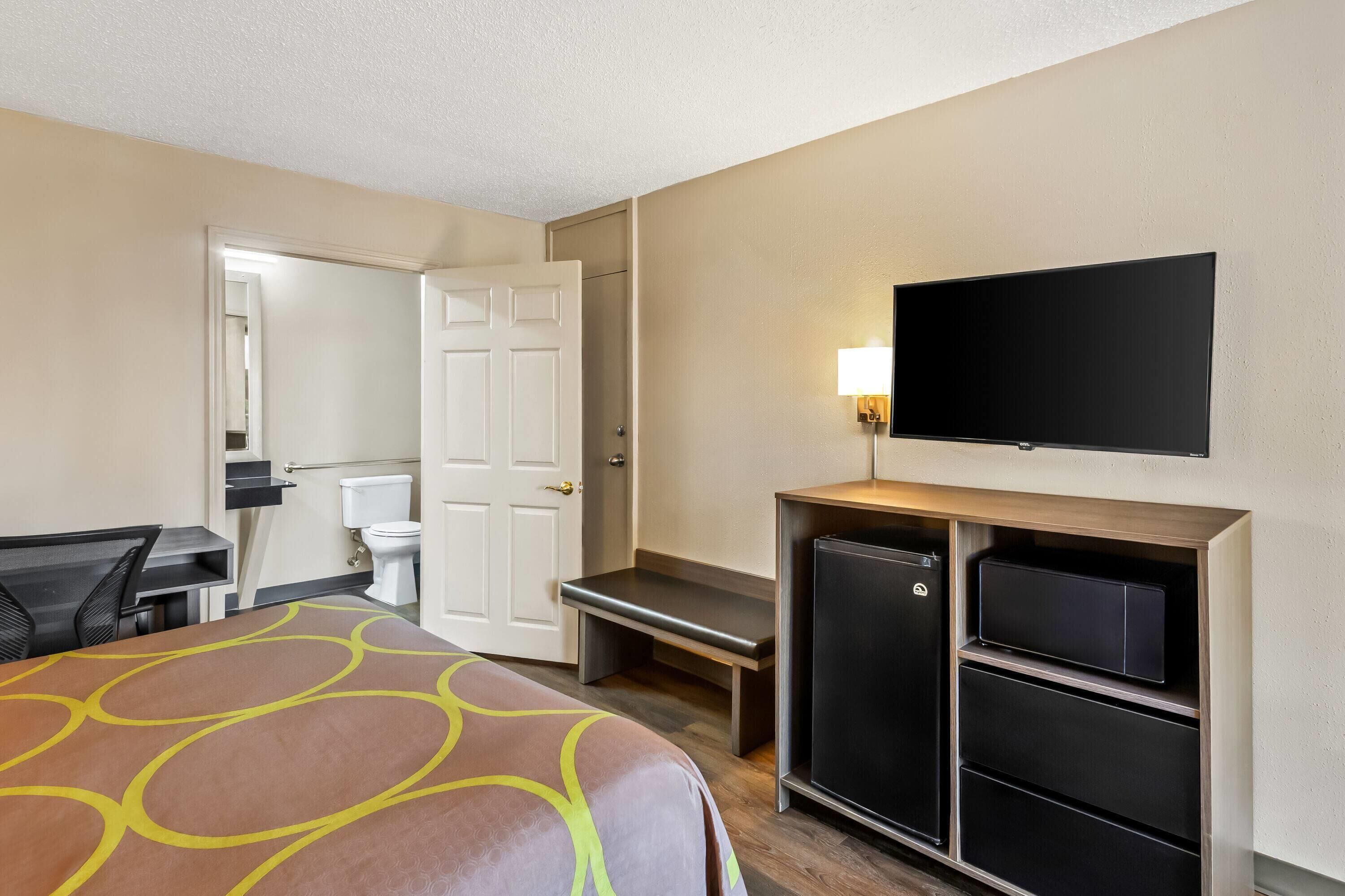 Una habitación de hotel con cama, escritorio, televisión montada en la pared, banca y un refrigerador pequeño.