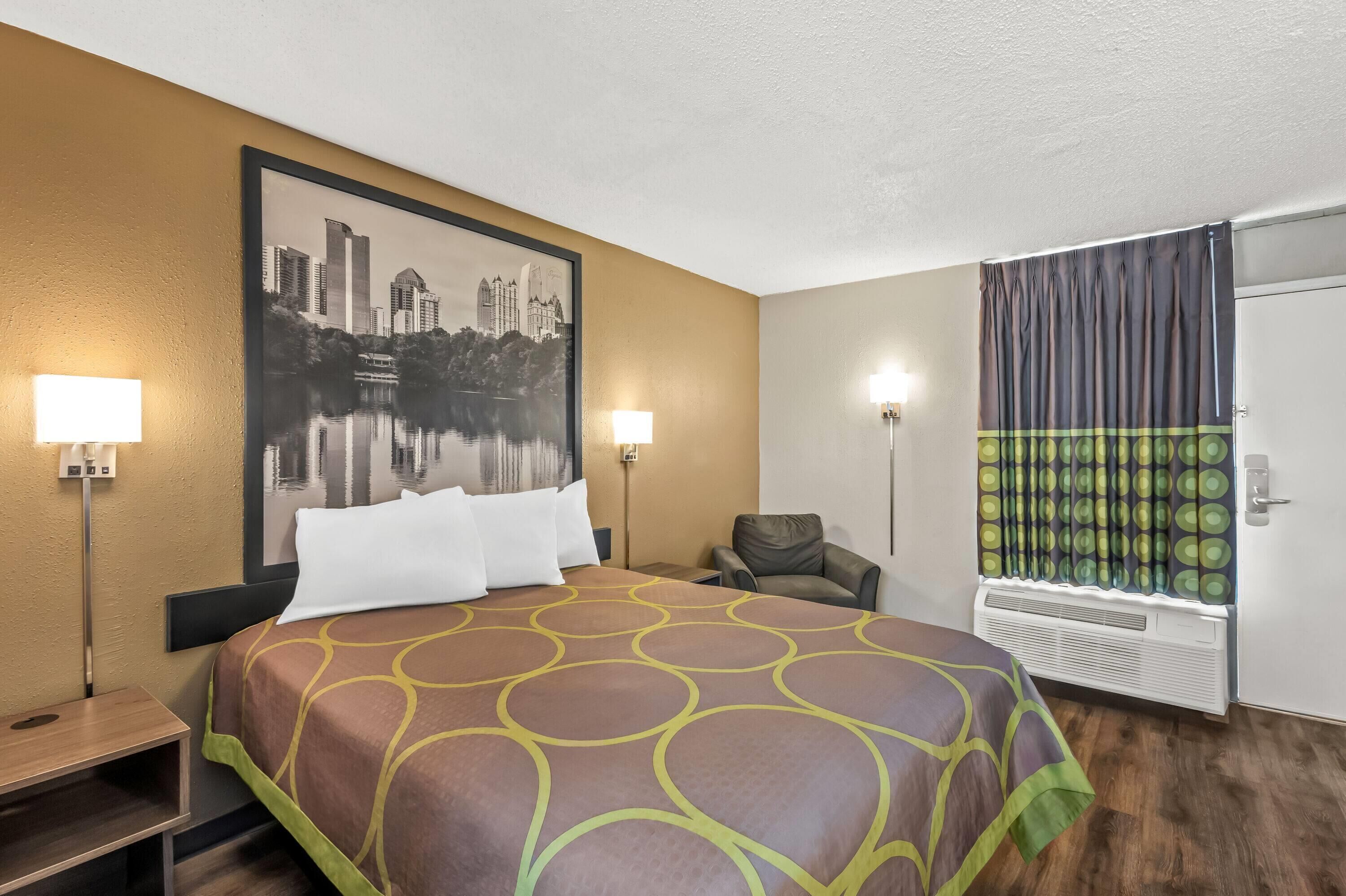 A hotel room with a large bed, two bedside lamps, a chair, a window with curtains, and a wall-mounted air conditioner.
