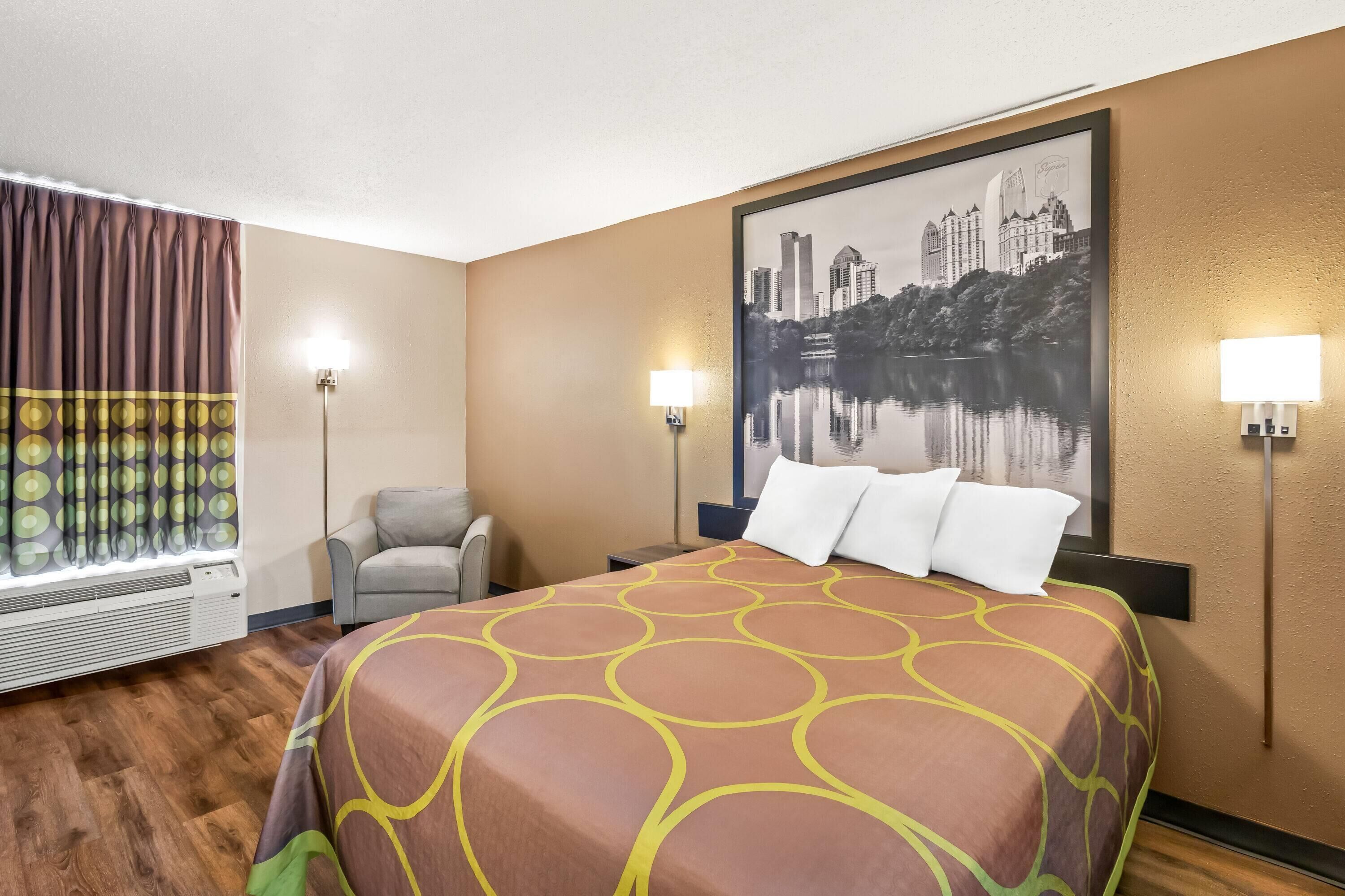 Una habitación de hotel con cama, una silla, una ventana con cortinas y un cuadro de un paisaje urbano en la pared.