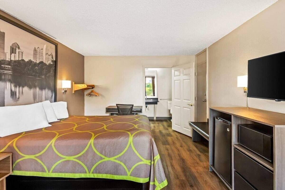 A hotel room with a bed, a desk, a chair, a TV, and a door leading to a bathroom.