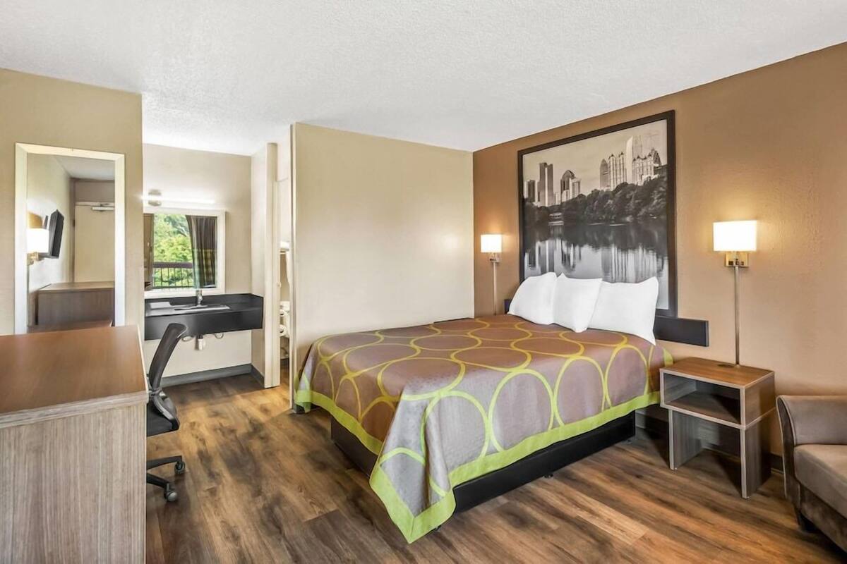 A hotel room with a bed, desk, chair, and a painting on the wall.