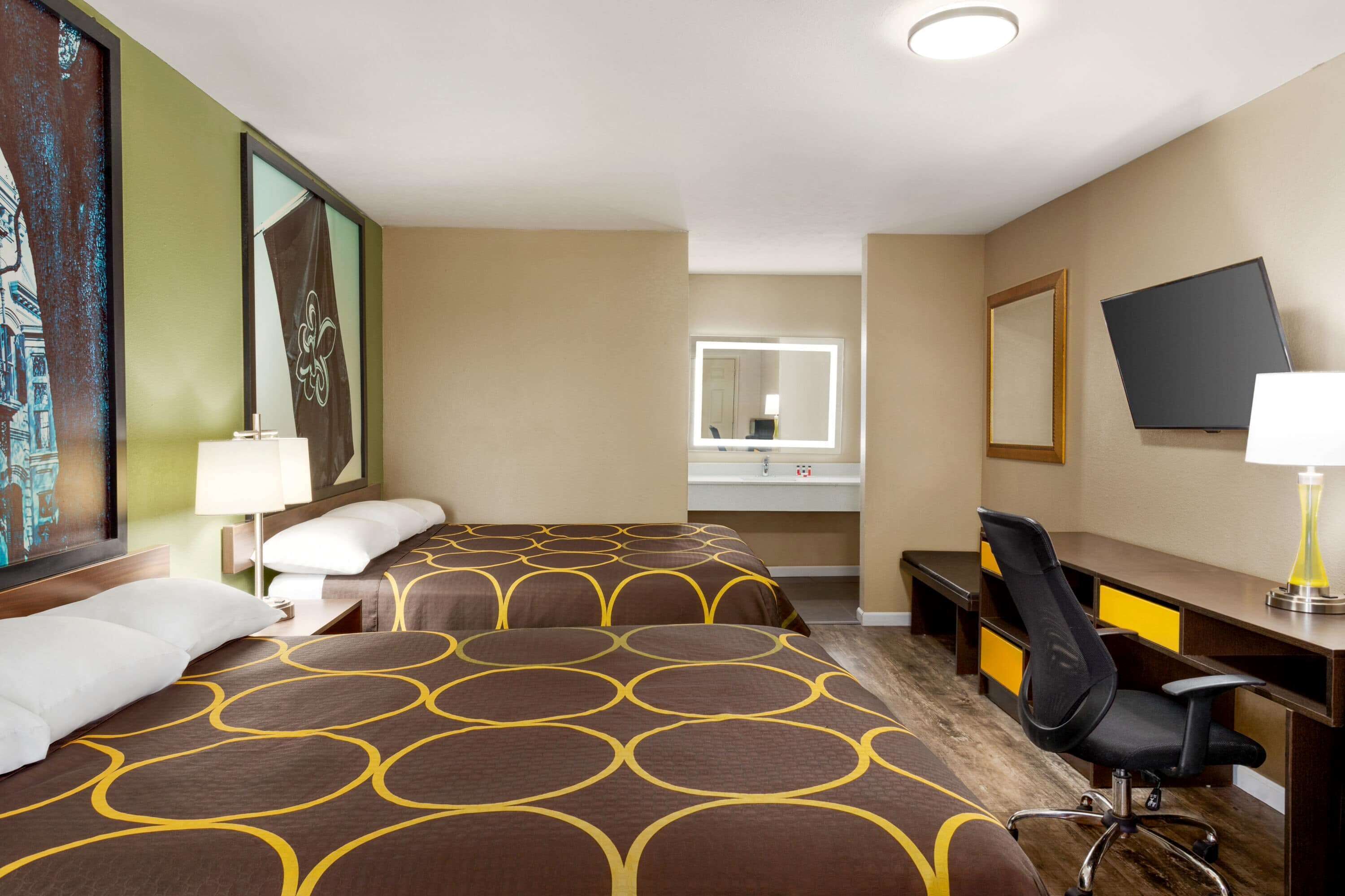 Ett hotellrum med två sängar, ett skrivbord, en stol, en TV och ett badrum som syns genom en glasdörr.