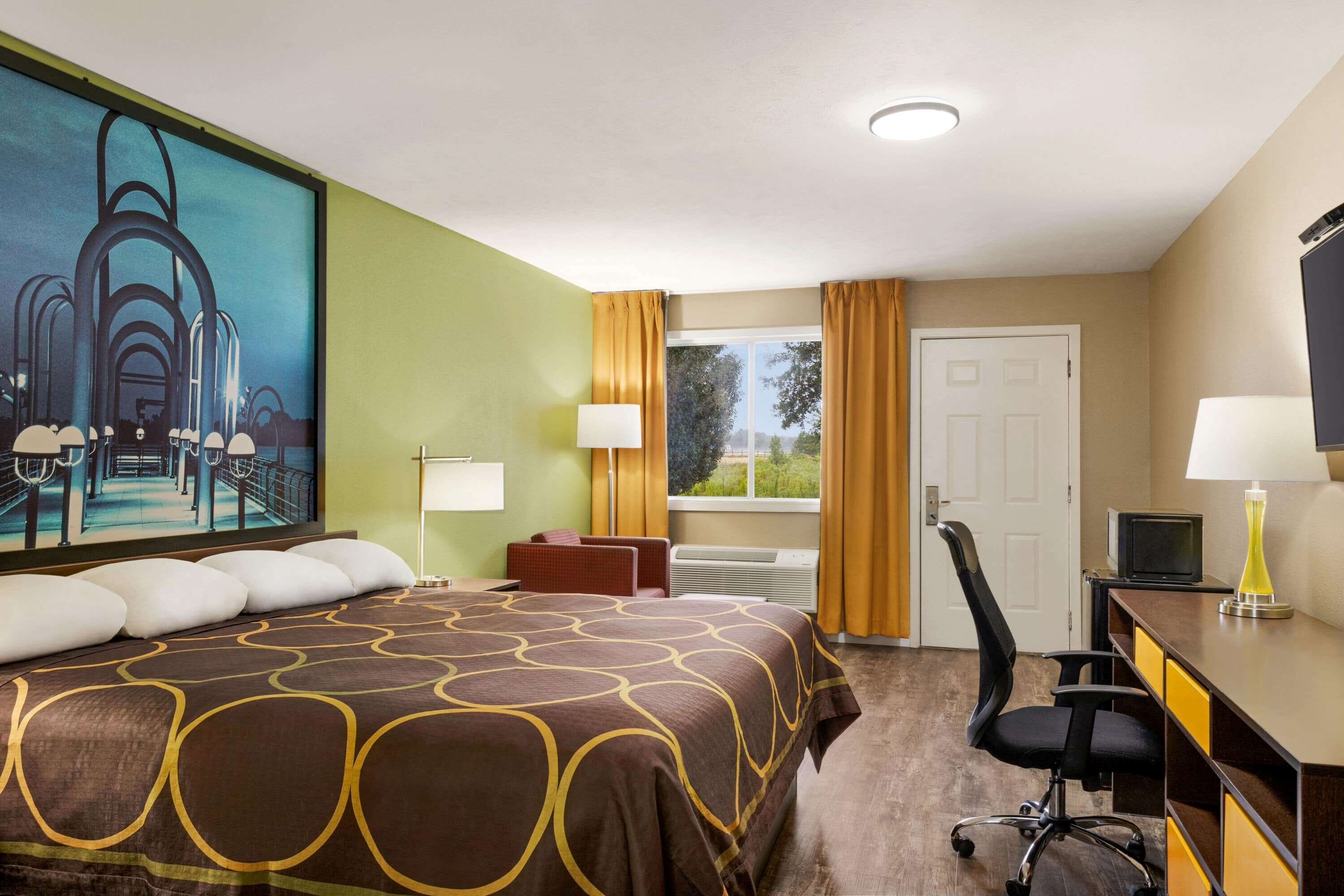 A hotel room with a large bed, a desk with a computer, a chair, a TV, and a window with curtains.