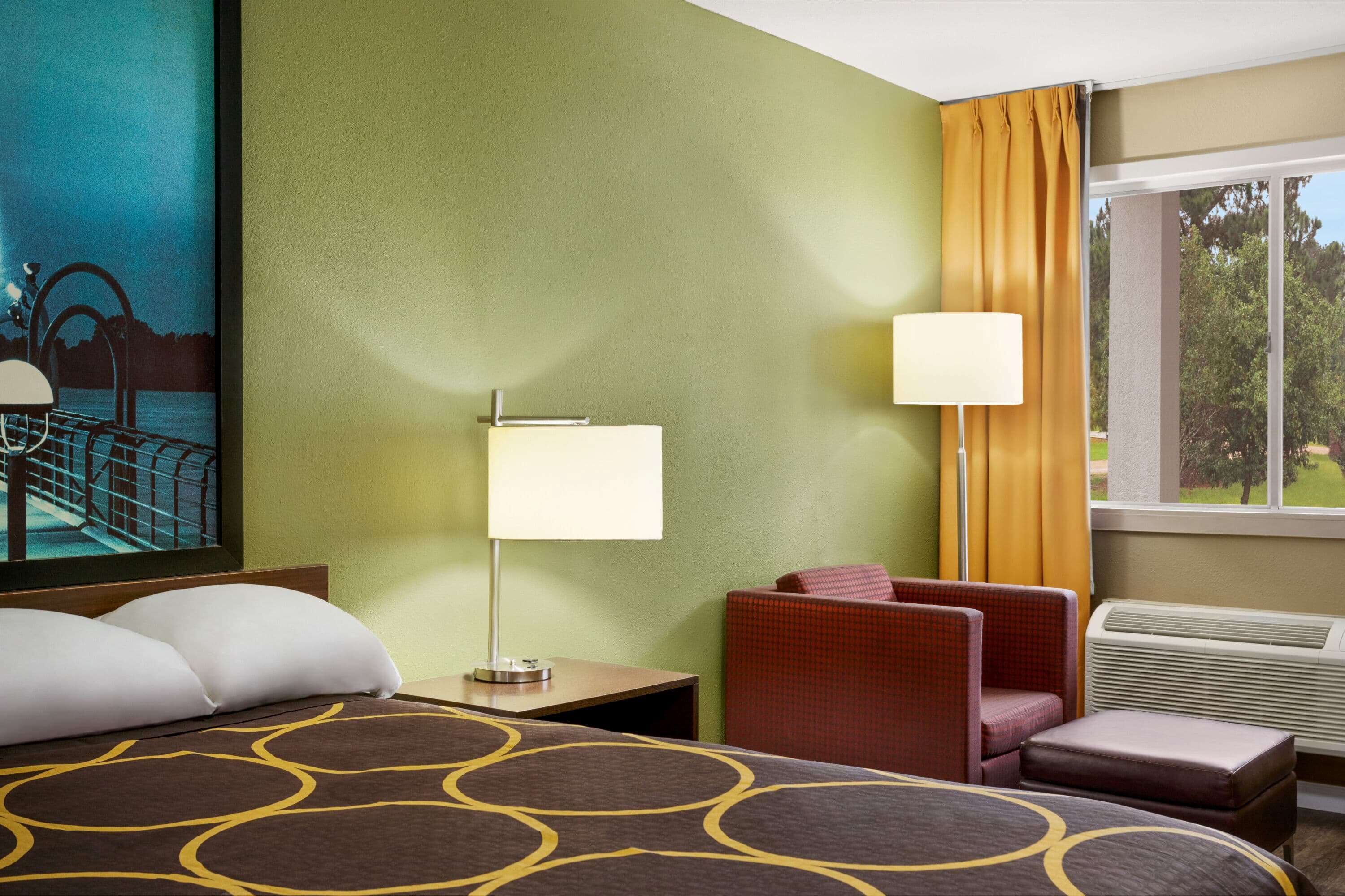 Habitación de hotel con una cama, dos mesitas de noche, una silla, un ventanal con cortinas y un cuadro en la pared.