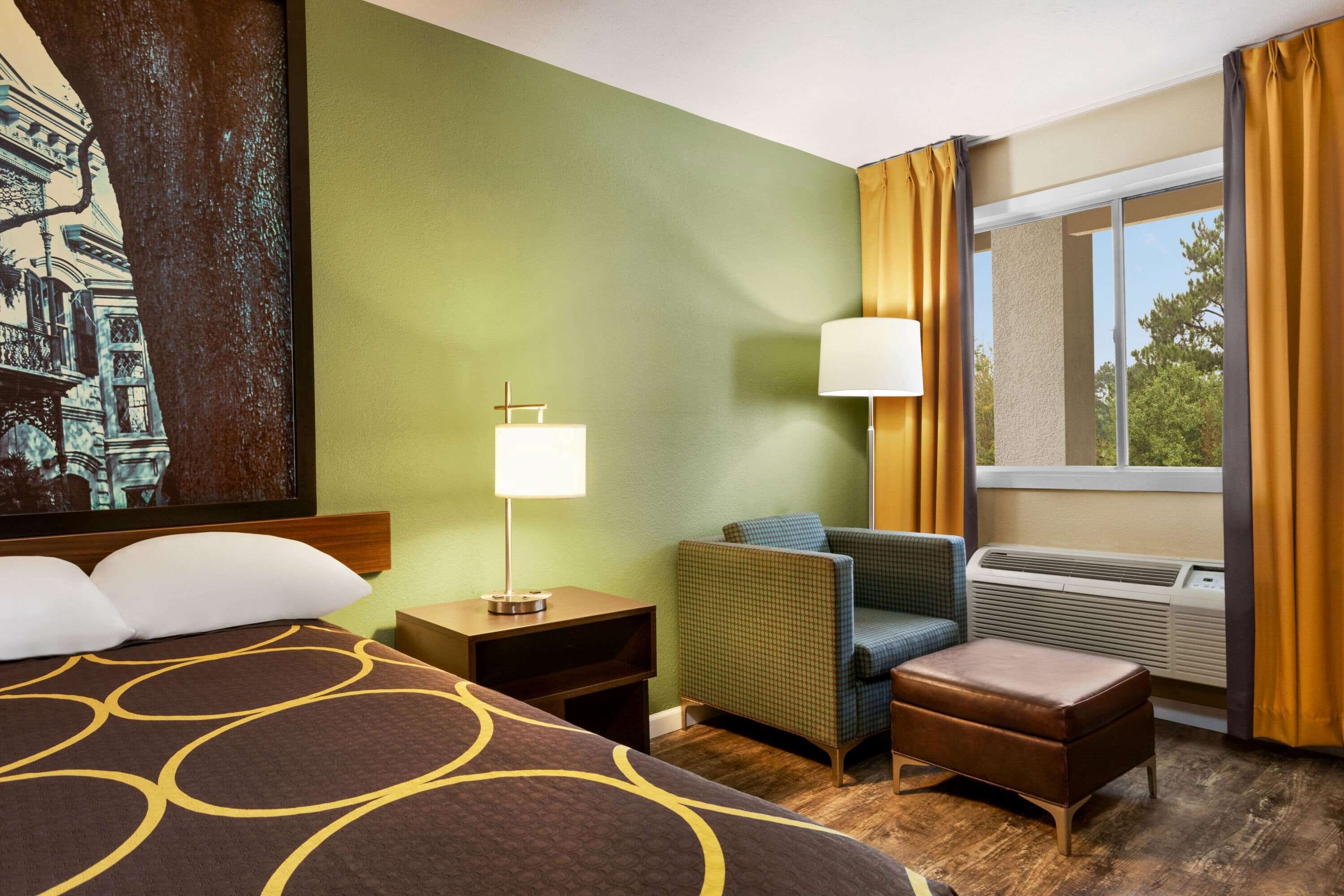 A hotel room with a bed, a chair, an ottoman, a nightstand, a lamp, and a large window with curtains.