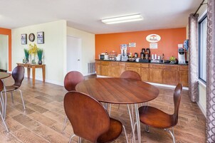Un comedor con mesas y sillas de madera, una encimera de cocina con equipo para preparar café, y un reloj en la pared.