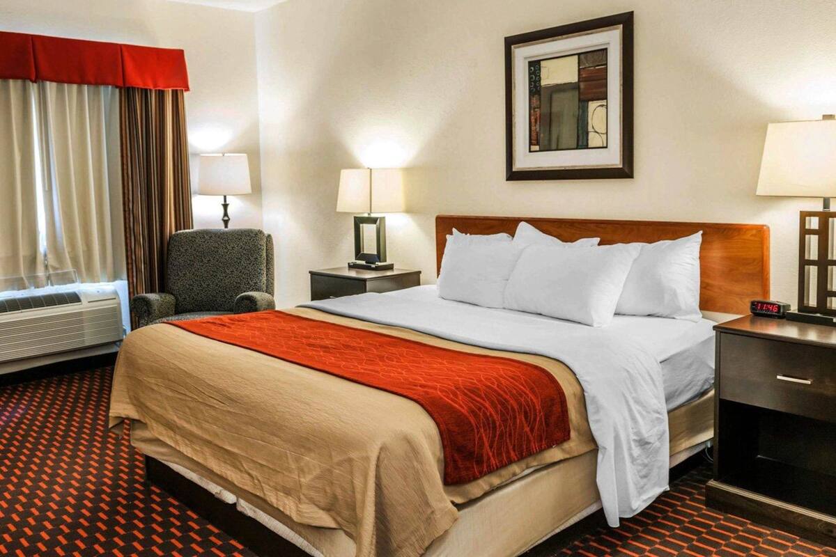 A hotel room with a bed, two bedside tables with lamps, a chair, a window with curtains, and a wall-mounted picture frame.