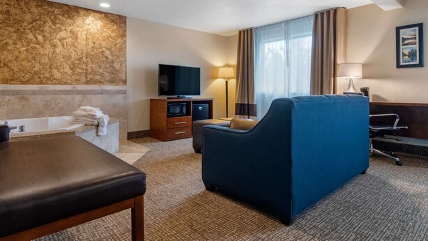 A hotel room with a massage table, a blue sofa, a flat-screen TV, a desk with a lamp, and a large window with curtains.