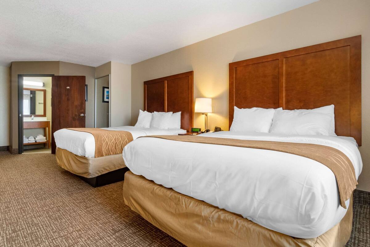 A hotel room with two beds, a wooden headboard, a bedside lamp, and a bathroom visible through an open door.