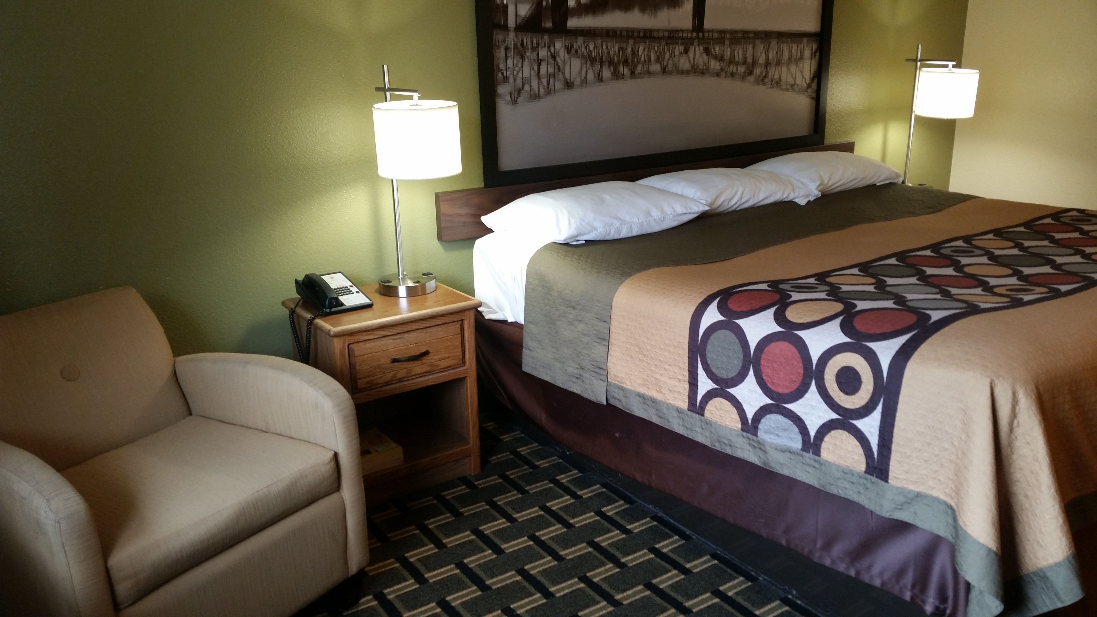 A hotel room with a bed, a bedside table with a lamp, a chair, and a patterned carpet.