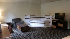 A modern bathroom with a freestanding bathtub, a grey armchair, a wooden bench, a television, and a patterned floor.