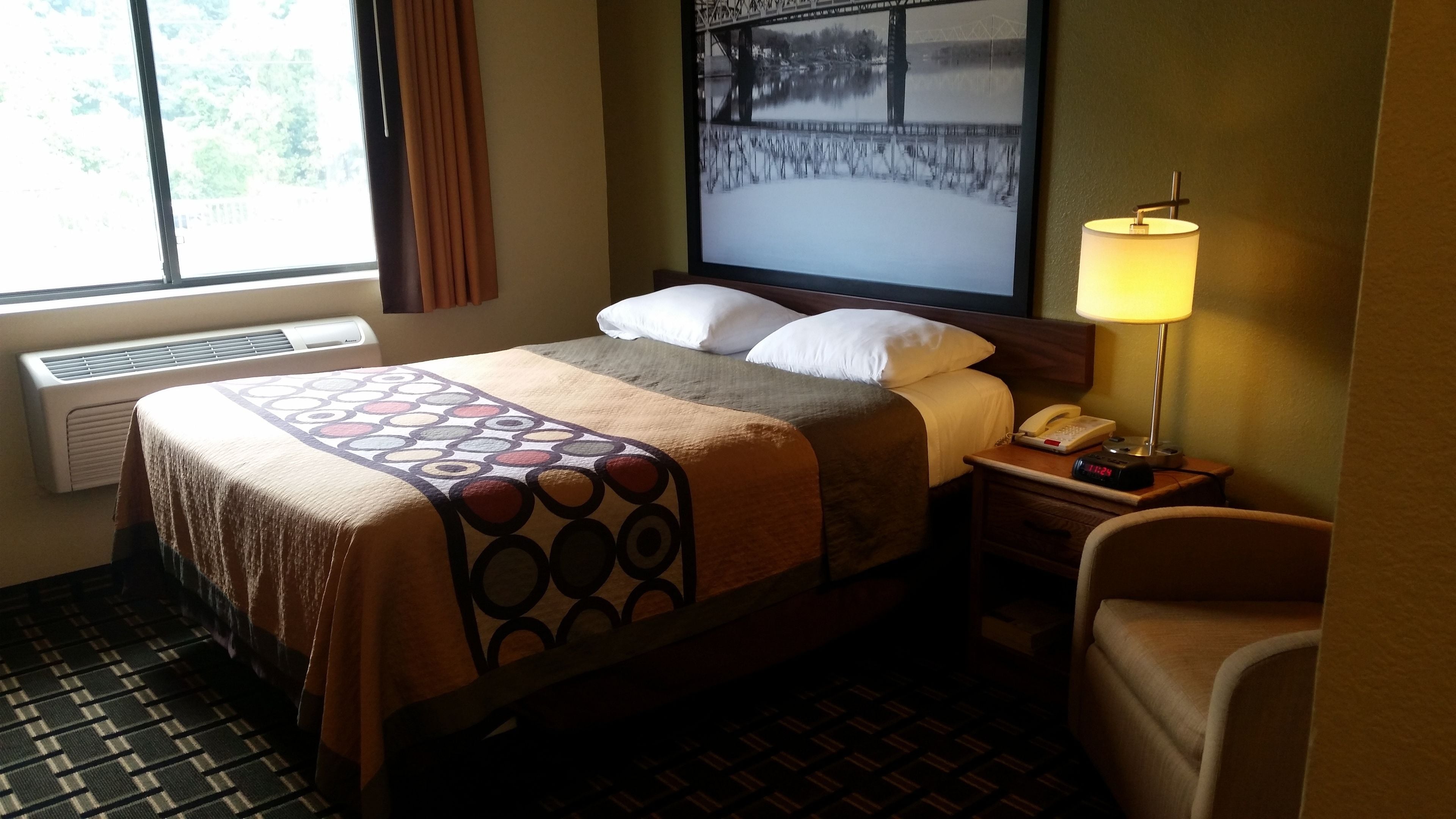 A hotel room with a bed, a nightstand with a lamp, a chair, a window with curtains, and a wall-mounted picture.