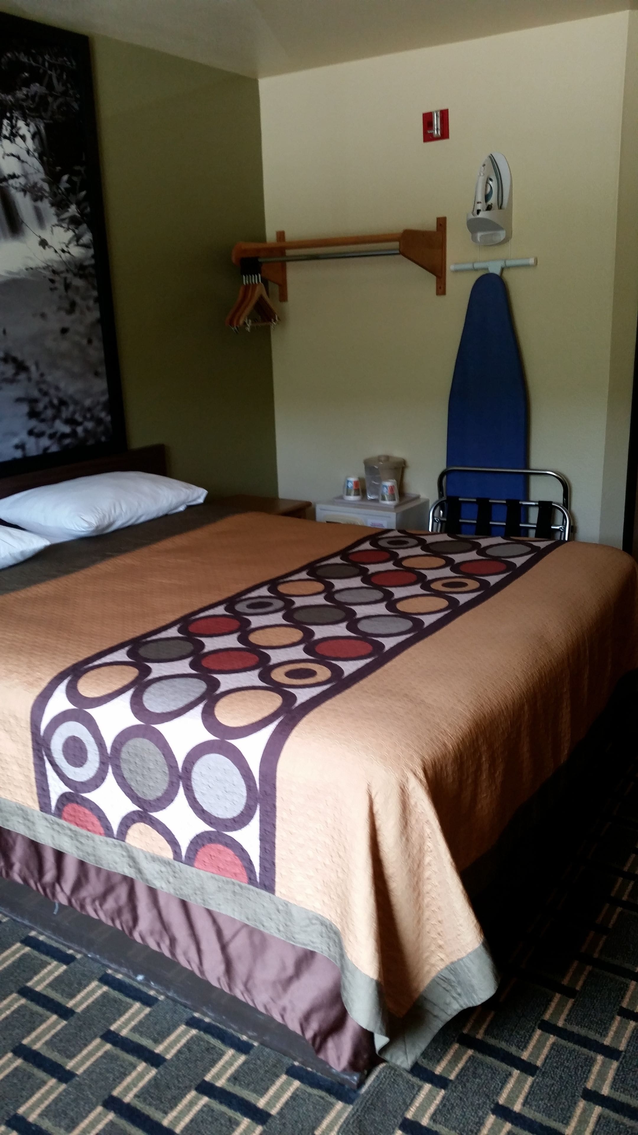 Een hotelkamer met een bed, een wandplank met een strijkplank, een klein tafeltje met een waterkoker en een schilderij aan de muur.