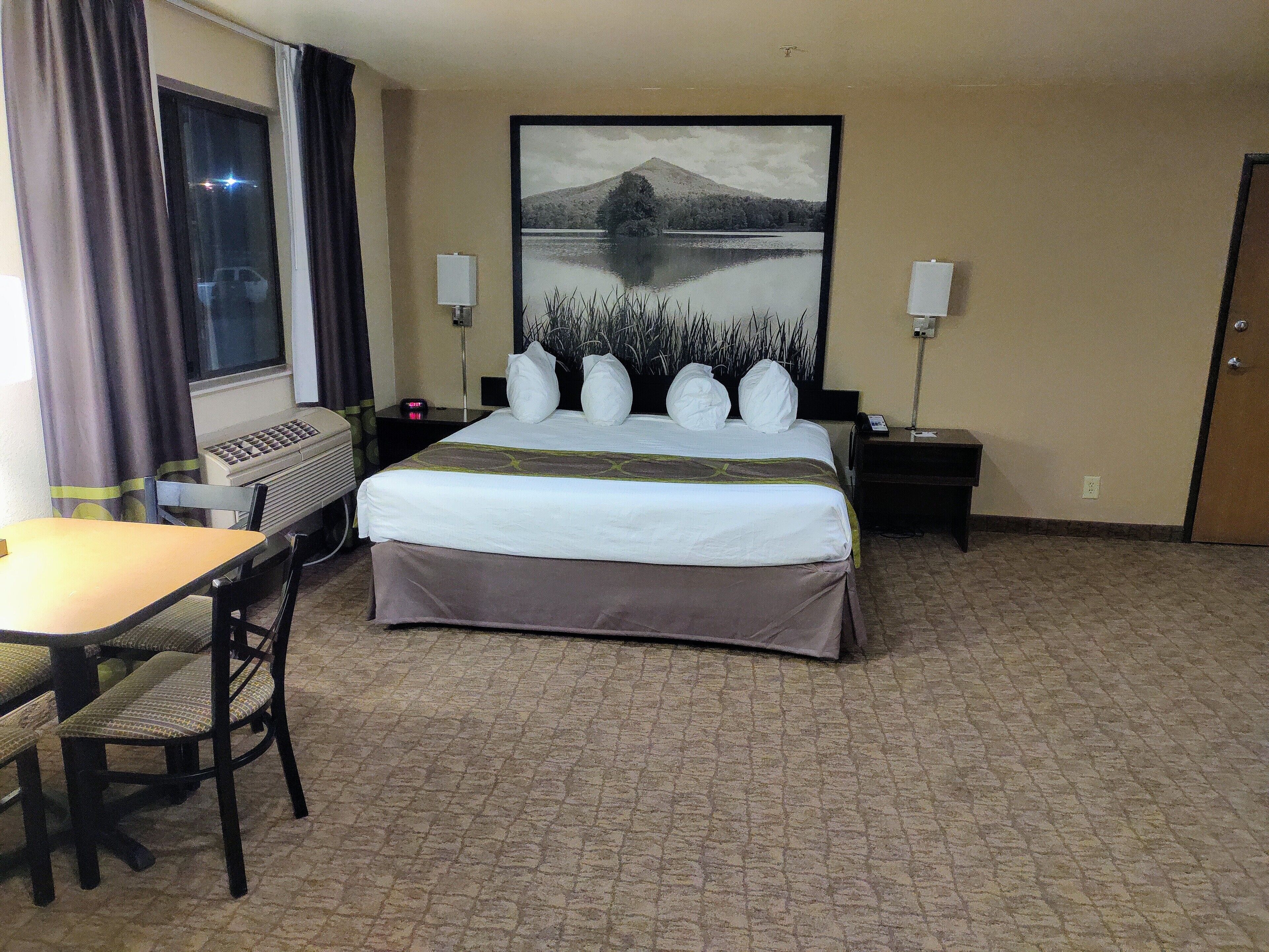 Ein Hotelzimmer mit Bett, Schreibtisch, Stuhl, einem Fenster mit Vorhängen, einem Bild an der Wand und einer Lampe auf dem Nachttisch.