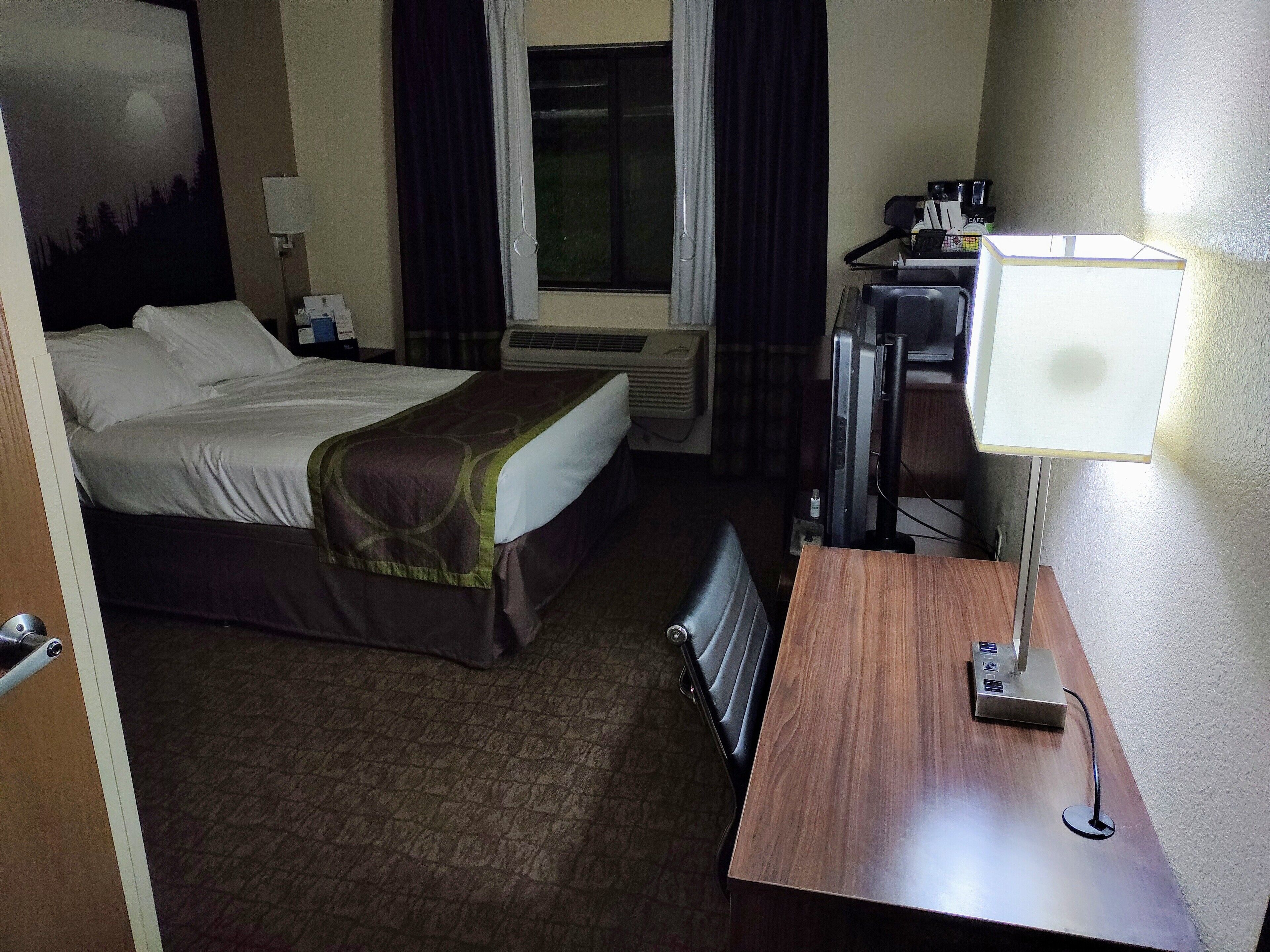 A hotel room with a bed, a desk with a lamp, an air conditioning unit, and a window with curtains.
