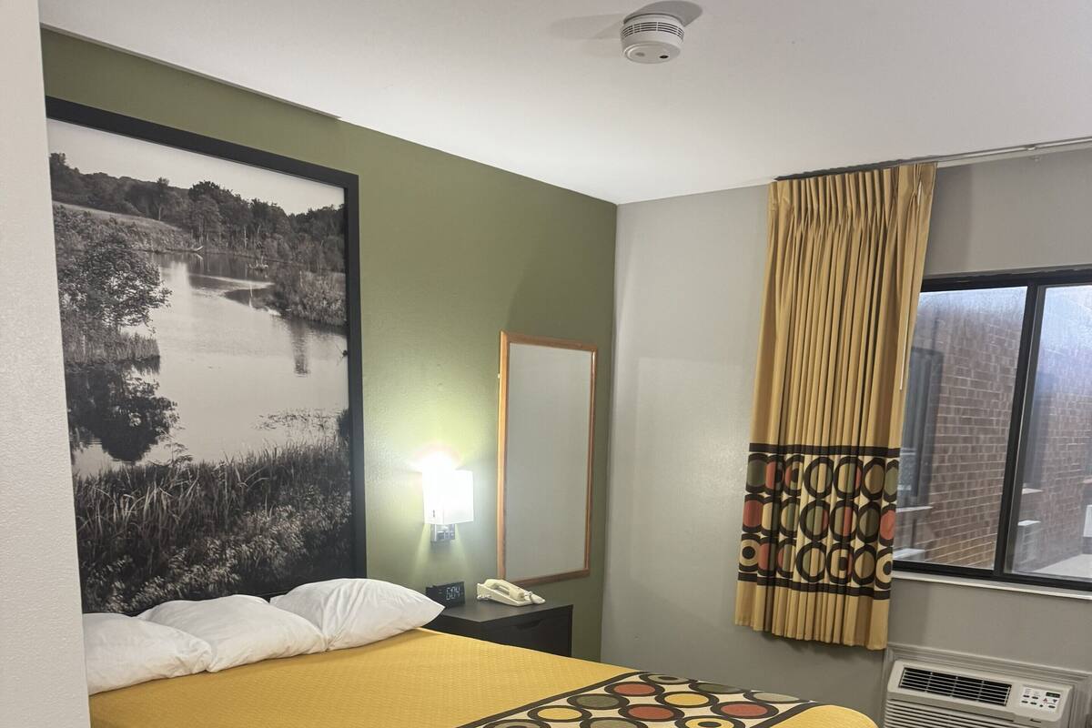 A hotel room with a bed, a nightstand, a lamp, a mirror, a window with curtains, and a radiator.