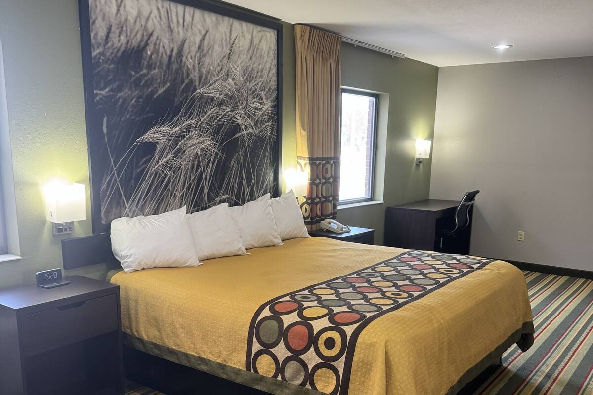 A hotel room with a bed, nightstands, a large wall art piece, a window with curtains, and a striped carpet.