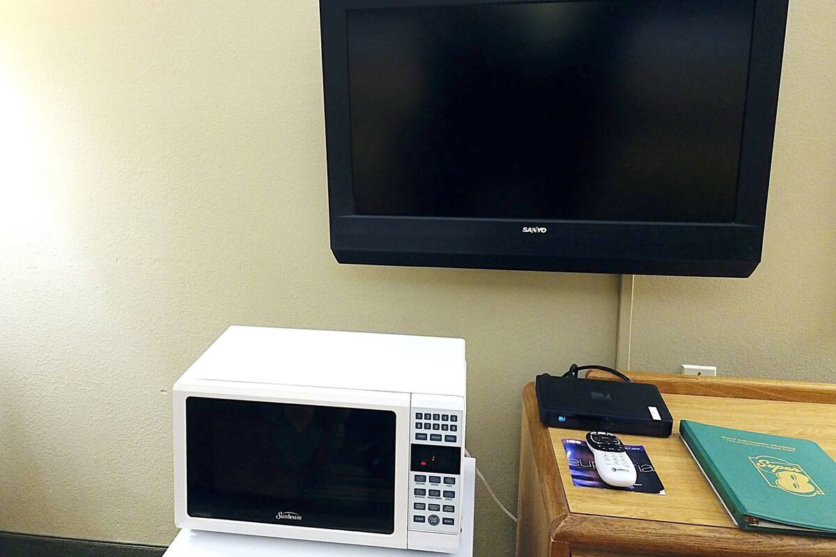 A room with a wall-mounted TV, a microwave, a wooden side table with a remote control, a book, and a power strip.