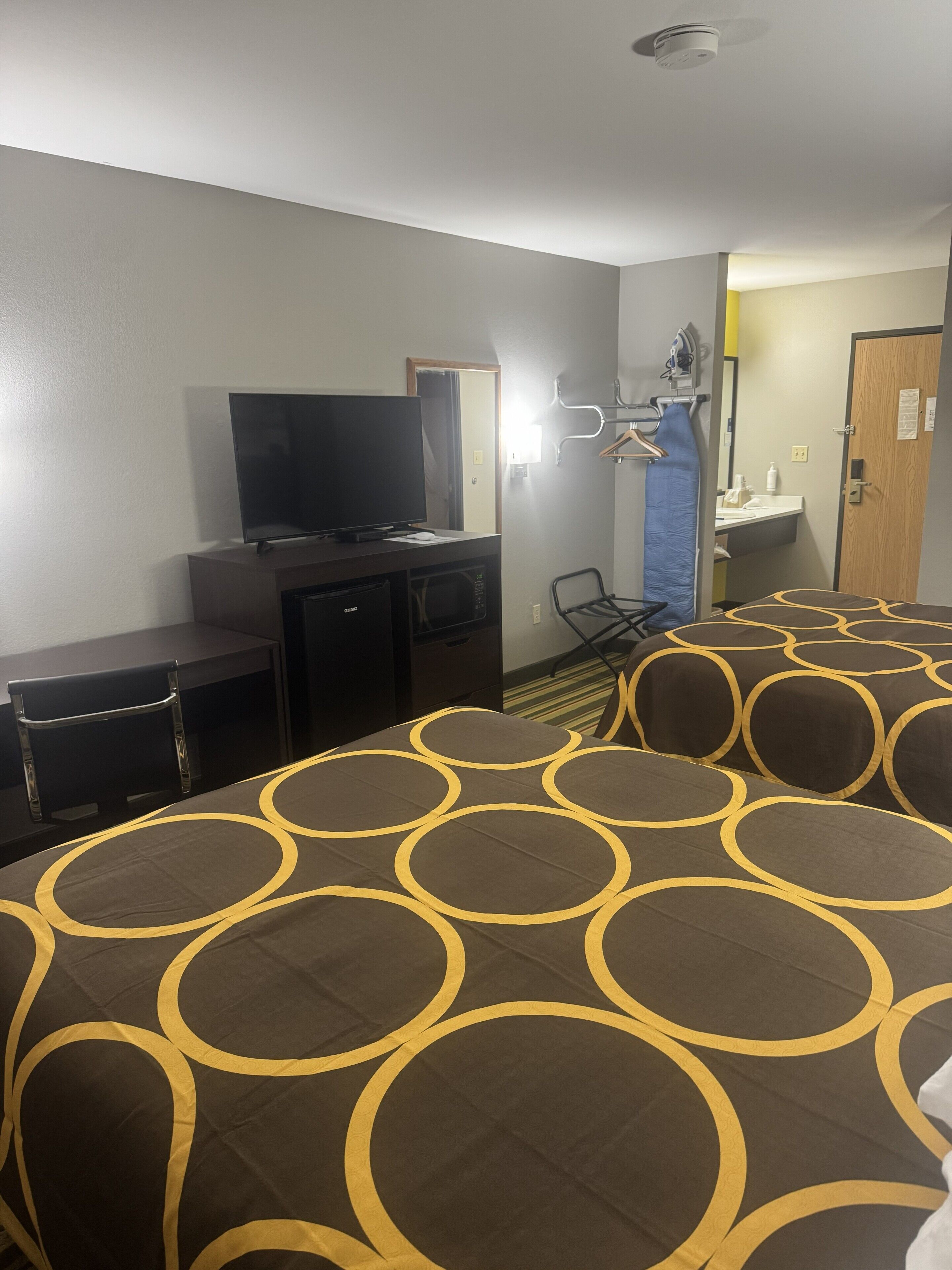 Una camera d'albergo con televisione, scrivania, sedia, letto con copripiumino decorato, specchio, lavandino e una porta.