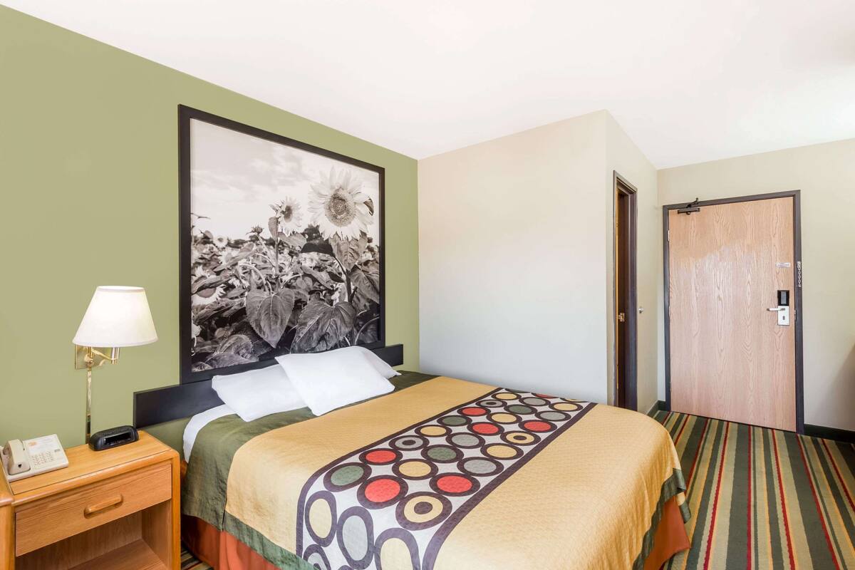 A hotel room with a bed, a nightstand with a lamp, a framed picture on the wall, and a door.