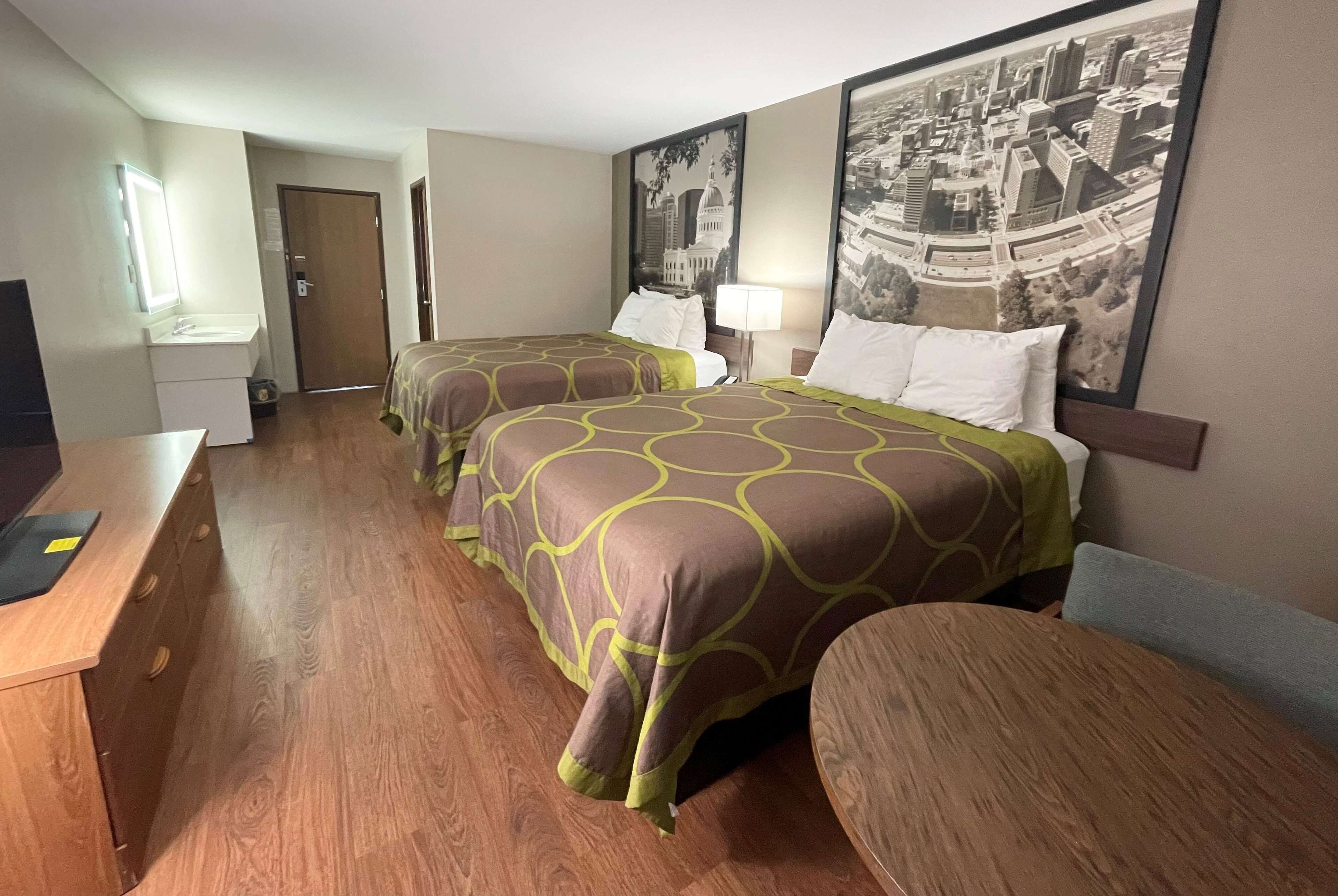 Quarto de hotel com duas camas, uma escrivaninha, uma cadeira, um abajur e uma pintura com vista para a cidade na parede.
