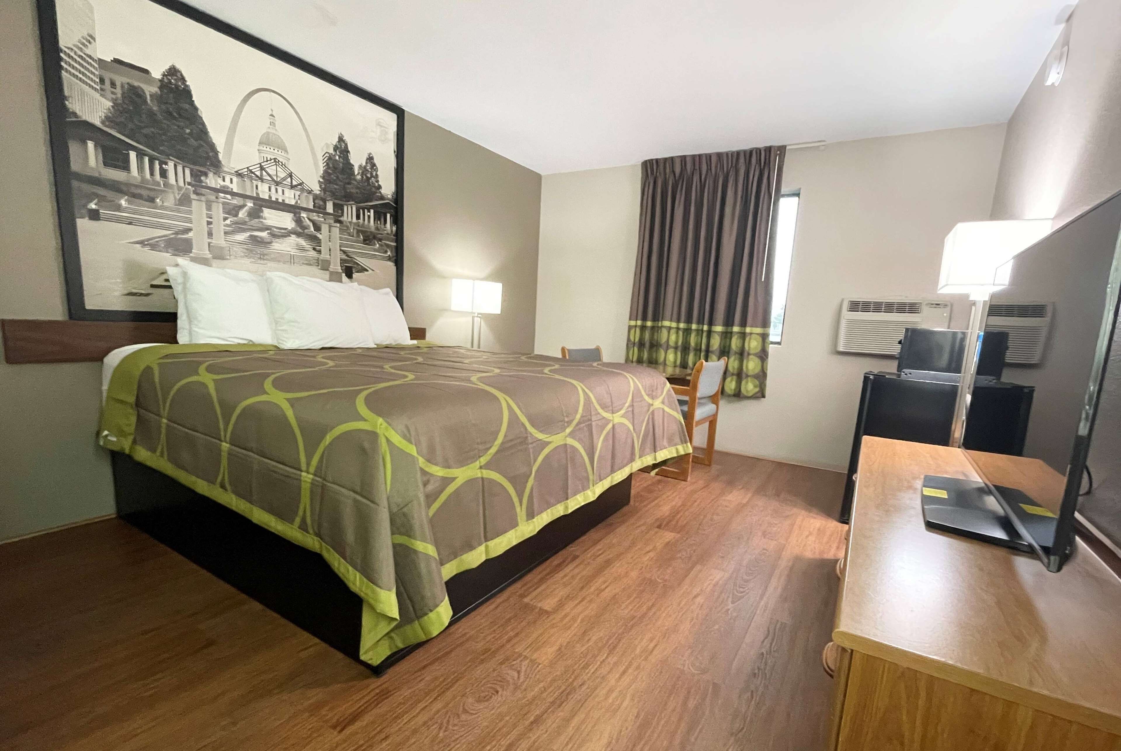 A hotel room with a bed, a desk, a chair, a lamp, and a large framed picture on the wall.