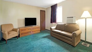 Hotelkamer met flatscreen televisie, een houten dressoir, een beige bank en een teal gekleurd tapijt, en een raam met gordijnen.