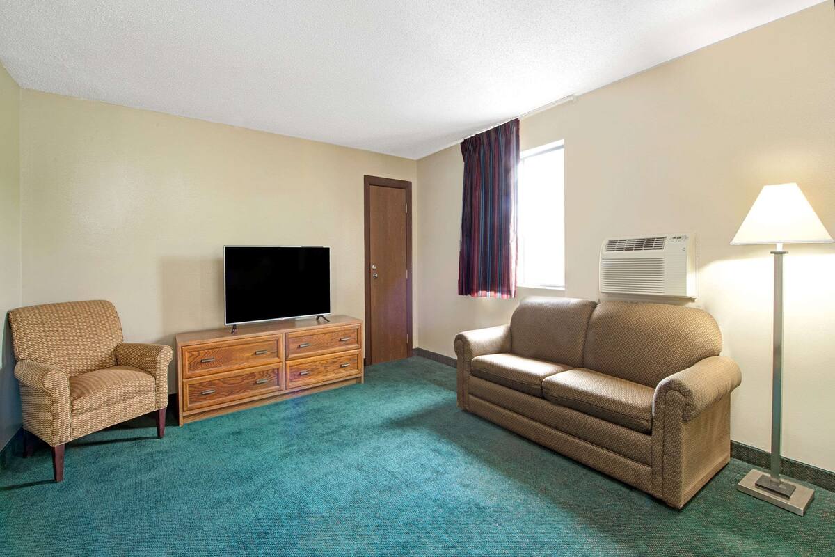 Une chambre d’hôtel équipée d’un téléviseur à écran plat, d’une commode en bois, d’un canapé beige, d’une moquette turquoise et d’une fenêtre avec des rideaux.