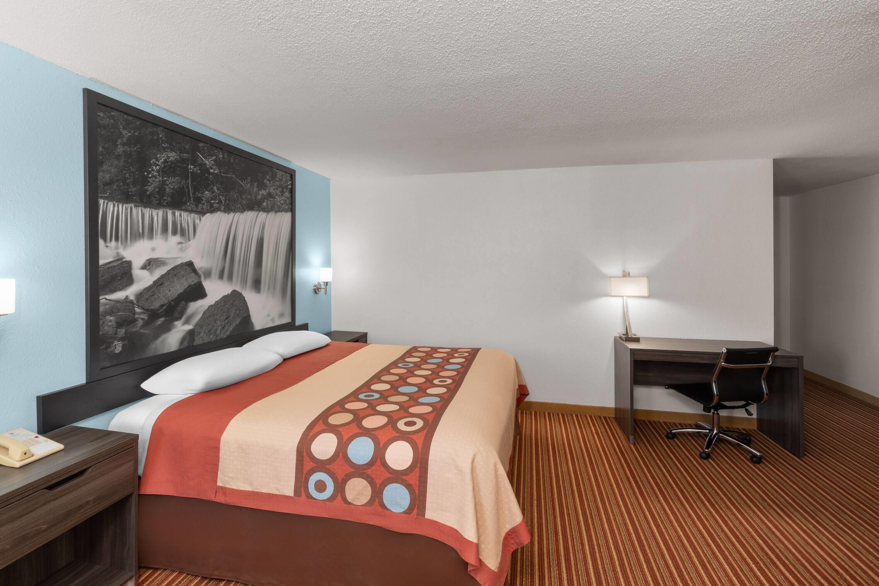 Quarto de hotel com cama, escrivaninha, cadeira, luminária e uma pintura grande na parede.