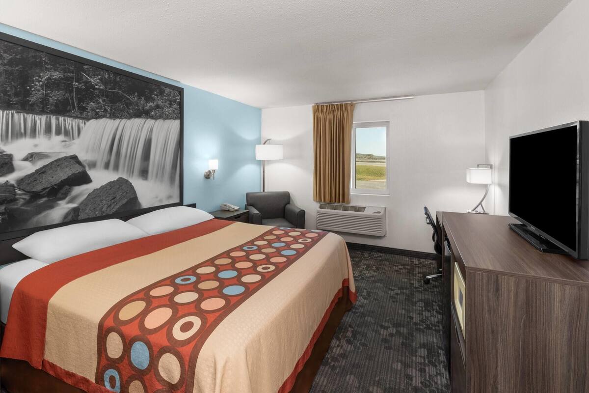 Une chambre d’hôtel équipée d’un lit, d’une télévision, d’un bureau, d’une chaise, d’une lampe, d’un climatiseur et d’une fenêtre avec des rideaux.