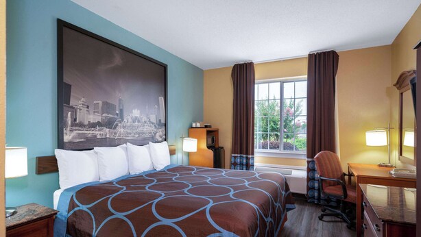 Ein Hotelzimmer mit einem großen Bett, einem Schreibtisch, einem Sessel, einem Fenster mit Vorhängen und einem Stadtbild an der Wand.