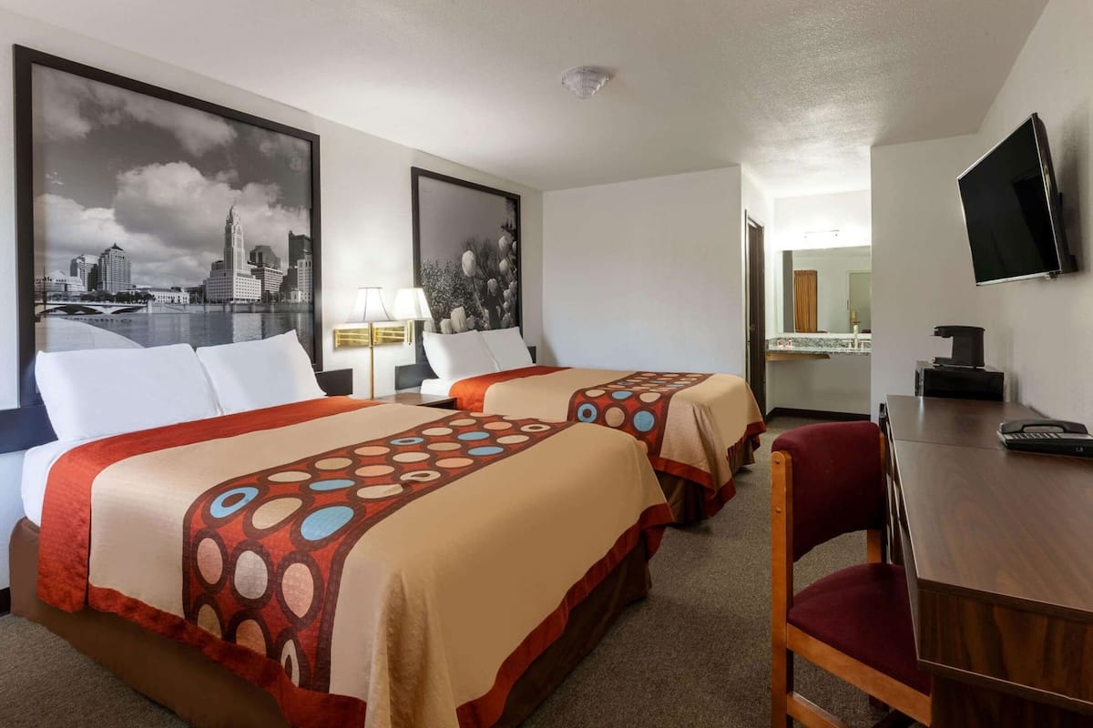 A hotel room with two beds, a desk, a chair, a TV, and artwork on the walls.