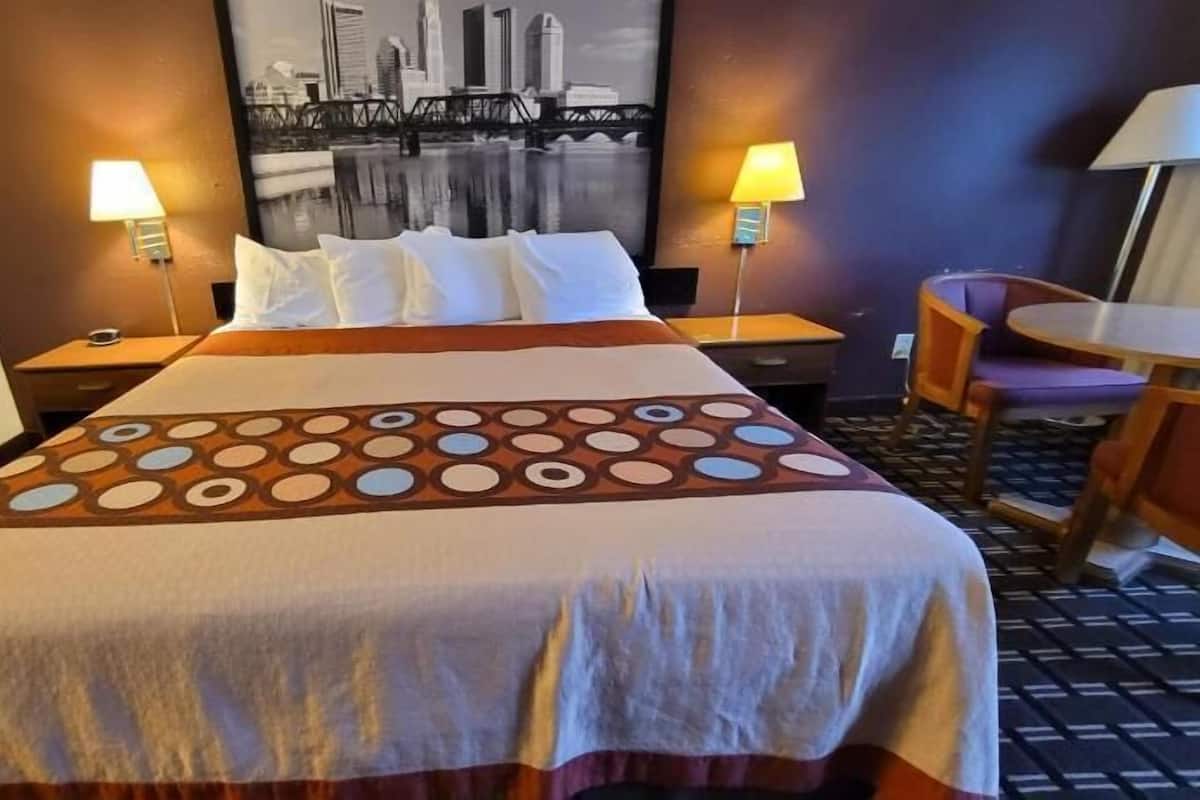 A hotel room with a large bed, two bedside lamps, a cityscape view, and a small table with chairs.
