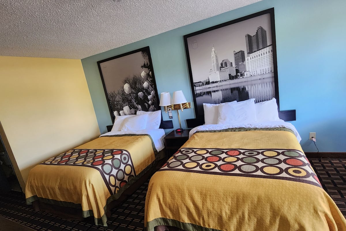 A hotel room with two beds, each with a patterned bedspread, a nightstand with a lamp, and a wall-mounted TV.